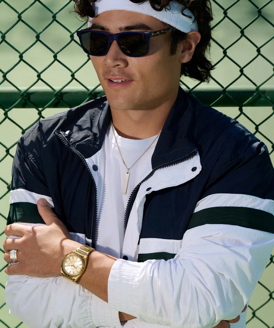 Shot of a man waiting his turn on the tennis court, wearing sunglasses, a gold Fossil necklace, a Machine Knurled Texture ring and an Everett Chronograph Gold-Tone Watch, all by Fossil.